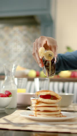 Vertical video: African American woman pouring syrup over pancakes at table making daughter laugh. Breakfast, family, joy, warmth, connection, candid, vertical video