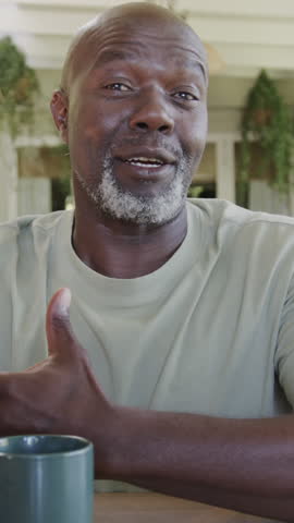 Vertical video: Senior African American man raising thumb, speaking for emphasis on porch with mug. Elderly, senior, contemplation, outdoor, candid, lifestyle, vertical video