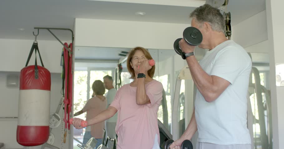 Entering home gym senior couple holding pink and black dumbbells curling arms in mirror for fitness. Fitness, strength, wellness, training, motivation, lifestyle, health