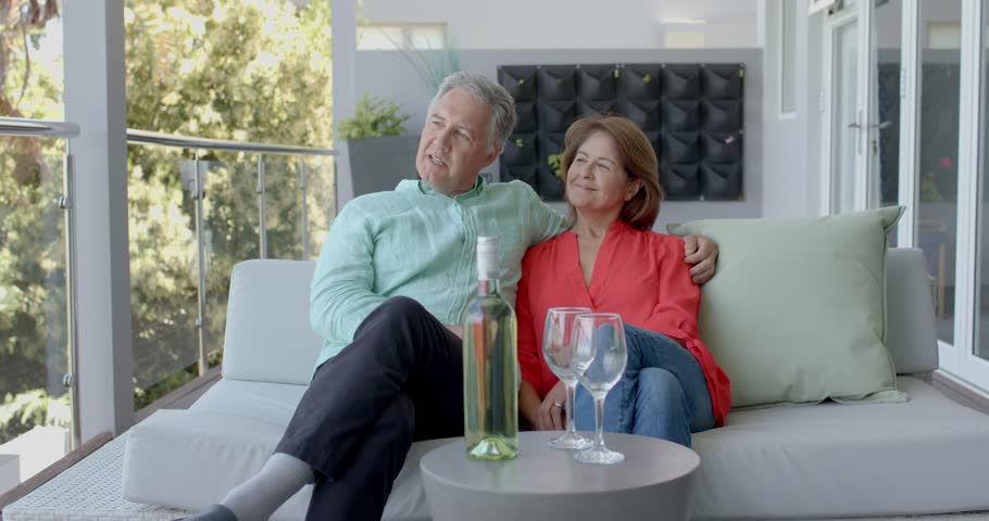 Senior couple smiling on balcony uncorking chilled bottle pouring wine into glasses toasting moment. Romantic, leisure, elegance, serenity, celebration, outdoor, lifestyle