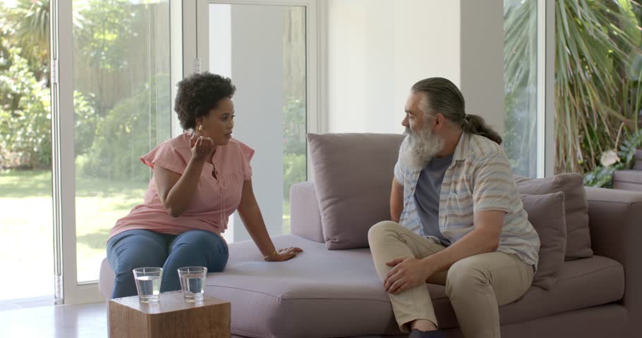 Diverse couple turning toward each other while gesturing and explaining on sofa near sliding door. Conversation, communication, domestic, lifestyle, modern, casual, interaction