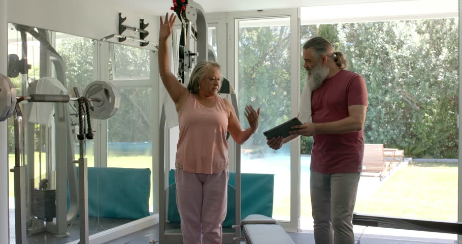 Fitness trainer cueing senior woman stretching overhead side forward on gym machine for flexibility. Geriatric fitness, flexibility, wellness, personal training, home gym, exercise routine, active