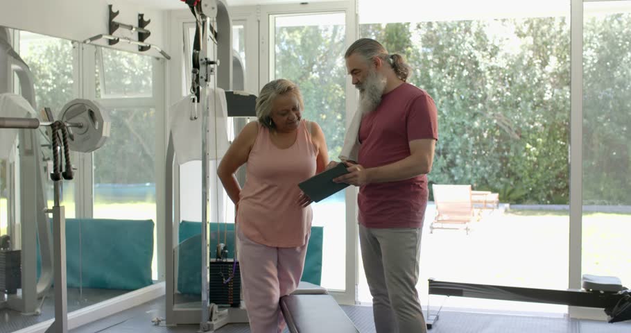 Male trainer tapping tablet explaining exercise plan senior woman nodding at cable machine home gym. Wellness, personal training, strength, aging, digital fitness, lifestyle, motivation