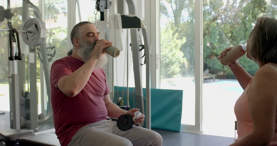 After drinking shakes, senior couple mirroring bicep curls with dumbbells in home gym for strength. Fitness, strength, healthy lifestyle, outdoor, domestic, wellness, determination