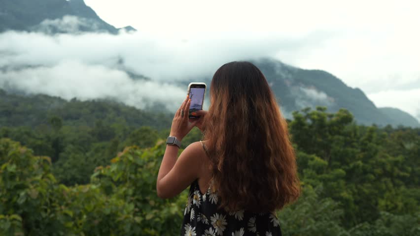 Young woman traveler taking photo of misty Doi Luang mountain with smartphone, Chiang Dao, Northern Thailand.