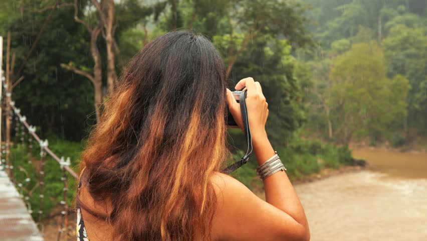 Young Woman Standing on Suspension Bridge Photographing River with Vintage Camera in Thailand