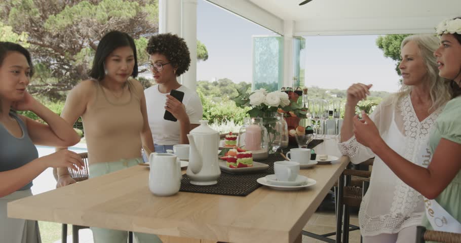 Diverse female friends picking up smartphone from wooden table edge and posing for selfie on patio. Friendship, celebration, leisure, outdoor, lifestyle, camaraderie, vibrant