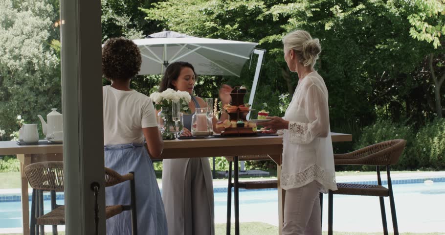 Reacting to senior woman handing cake, Chinese hostess arranging vases pitchers glasses by pool. Resort, leisure, elegance, celebration, outdoor, hospitality, sophistication