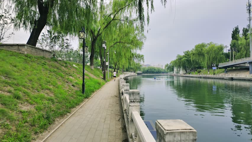 It’s a bit cloudy in the evening while walking by the river. The grass is lush green, and the river water is also green. Some people are jogging, and others are paddleboarding in the river.