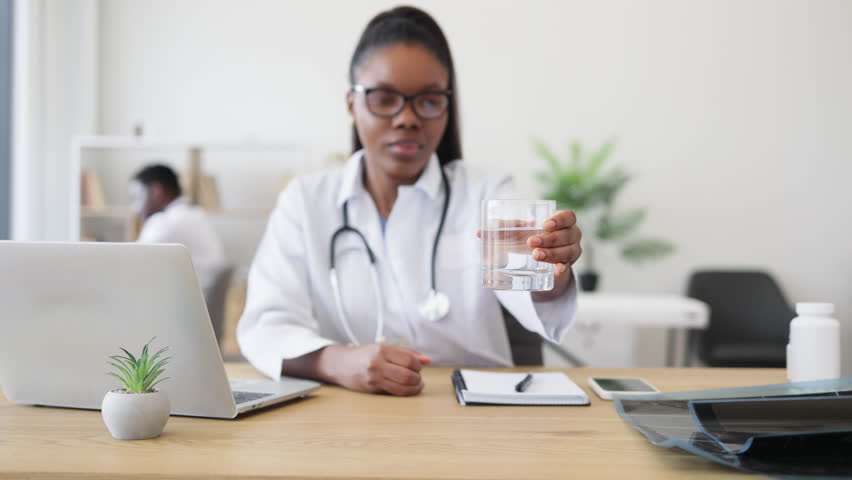 Young female doctor wearing glasses and lab coat explains benefits of clean water hydration. Office environment with colleague visible. Promotes health and healthy lifestyle emphasizing water