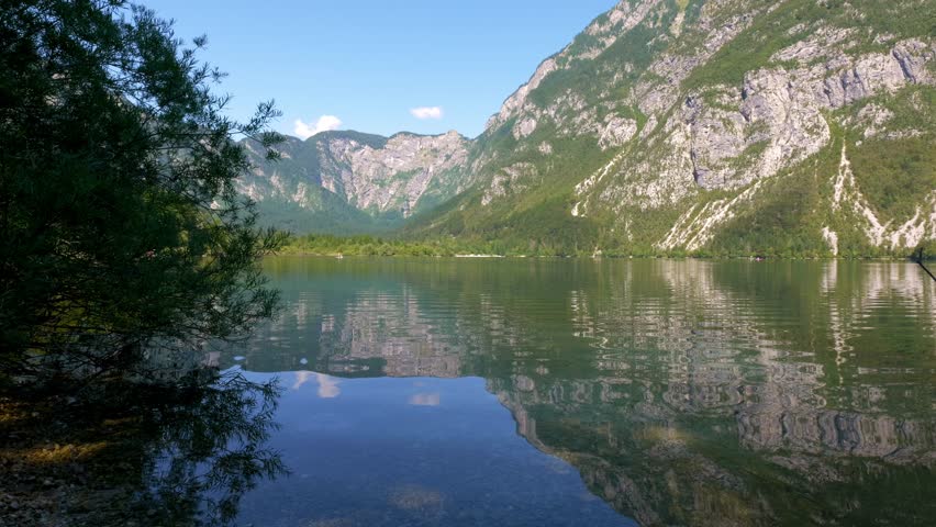 Calm lake water surface with blue sky reflection and the mountains. Lake Bohinj in Slovenia on a sunny morning. Peaceful natural surroundings with lake, mountains and the trees. Paradise in the Alps.