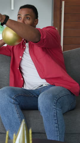 Vertical video: Mid adult African American man inflating gold balloon for party on gray sofa. Celebration, party, happiness, joy, festive, recreational, vertical video