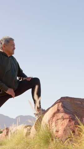 Vertical video: Senior man stepping onto red rock under mountains for lunge stretch, adjusting pose. Wellness, flexibility, outdoor, fitness, vitality, nature, vertical video