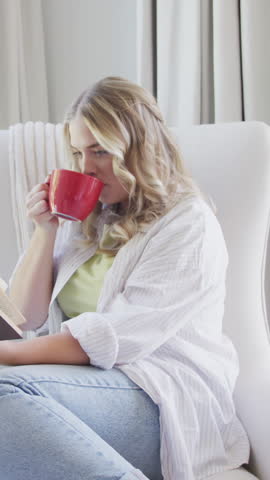 Vertical video: Woman settling into armchair at home sipping from red mug to focus on reading book. Cozy, comfort, relaxation, leisure, lifestyle, ambient, vertical video