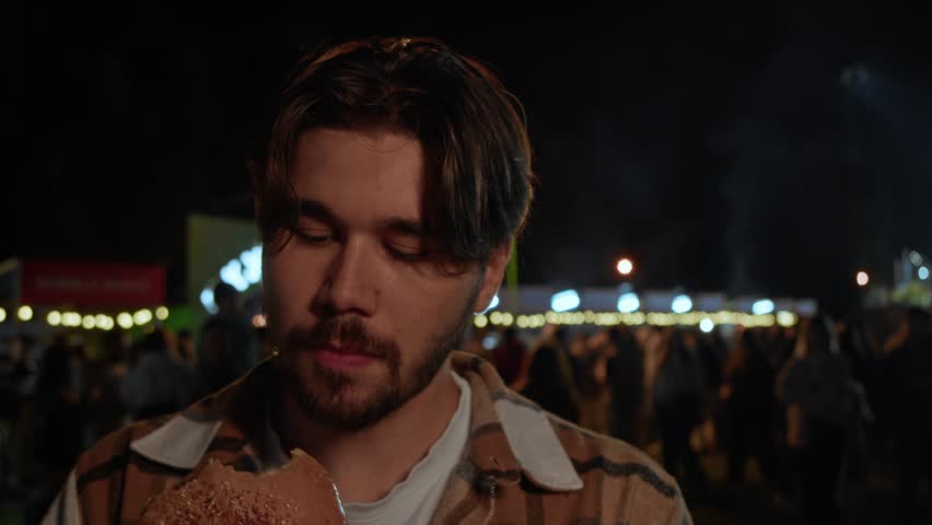 Young man in checked jacket enjoys large burger, taking big bite with gloved hands against lively night street food festival backdrop filled with lights and people. Portrait.