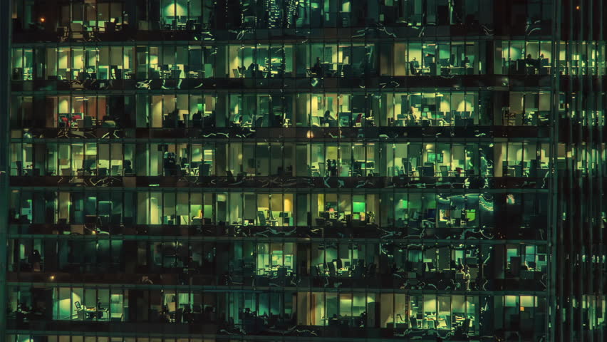 A vibrant city office building lit up at night, showcasing numerous workers through its bright, illuminated windows