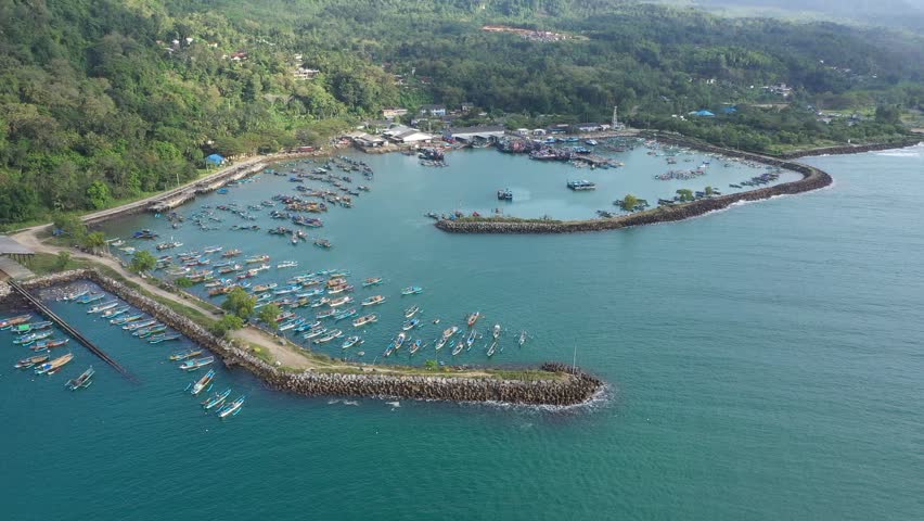 An aerial view of a bustling fishing port with numerous colorful boats moored in a calm harbor. The shot captures the coastal town nestled between green hills and the ocean.