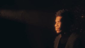 Young African American man in fluffy vest standing in beam of golden light illuminating face against dark background. Side profile, copy space - Powered by Shutterstock - Get 15% off with code: PIKWIZARD15