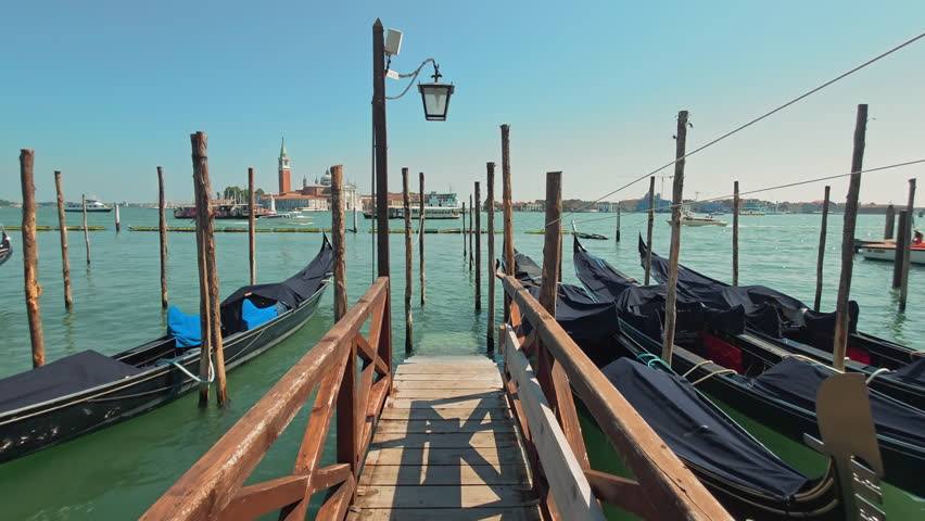 Wooden pier with moored gondolas in Venice. Empty gondolas covered and tied to wooden poles at a quiet pier in Venice, overlooking the calm lagoon on a sunny day.