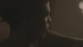 Shirtless Black man sitting in dimly lit room with soft warm light and looking away contemplatively. Close-up view - Powered by Shutterstock - Get 15% off with code: PIKWIZARD15