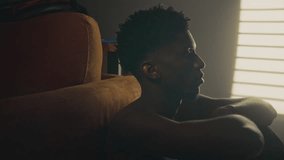 Young African American man with bare torso sitting alone in warmly lit bedroom with arms resting on knees, tilting his head beach and gazing upward pensively - Powered by Shutterstock - Get 15% off with code: PIKWIZARD15