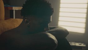 Portrait of young Black man sitting shirtless on floor with arms resting on knees and looking at camera, illuminated by soft light from window with blinds in cozy bedroom - Powered by Shutterstock - Get 15% off with code: PIKWIZARD15