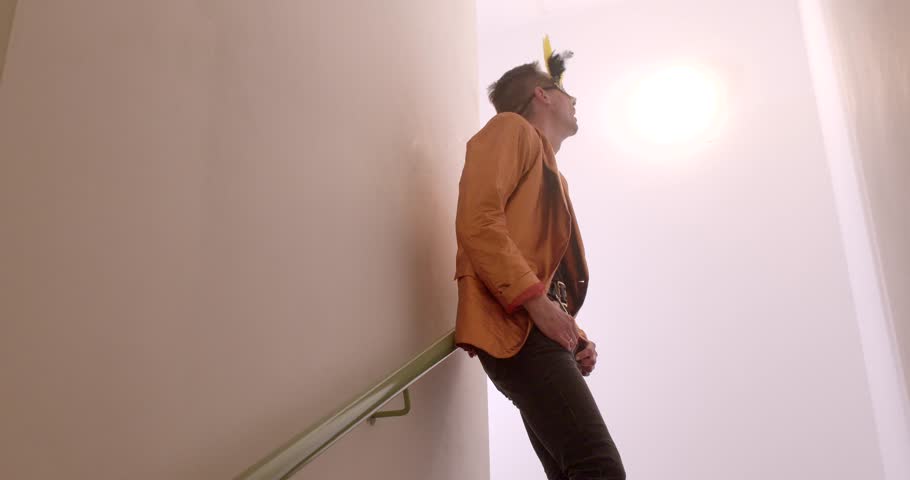 A stylish man in an orange jacket poses on a staircase, exuding a unique fashion sense. His playful demeanor and outfit create a captivating scene.