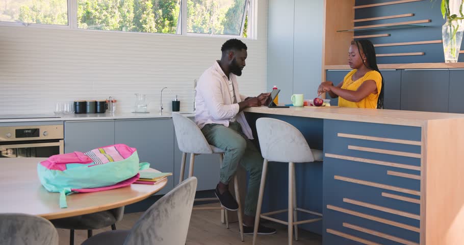 African American family tapping tablet slicing apples in kitchen while daughter arranging backpack. Family, domestic, modern, lifestyle, education, bonding, casual