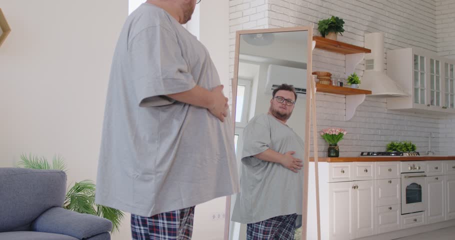 Overweight man checks belly reflection at home. He stands by a mirror in a bright kitchen, weighing a diet and healthier lifestyle to reduce obesity. Self-care and weight loss motivation.