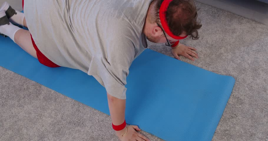 Overweight man does workout pushups indoors at home. Kneeling on a mat, he attempts modified exercise to build strength and mobility with proper form. Funny fitness training at home today.