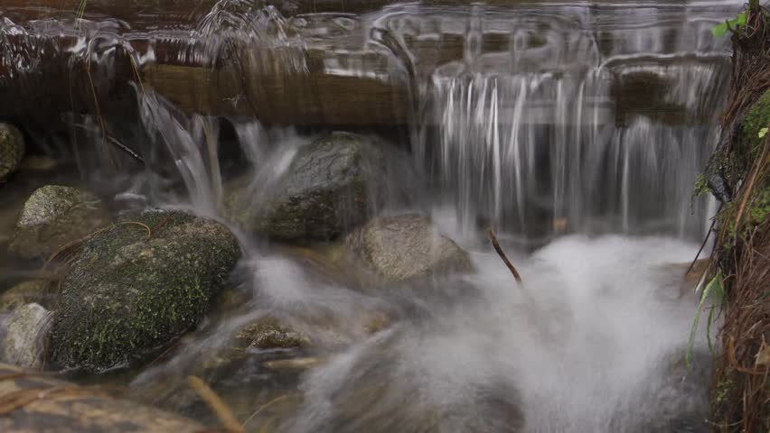 A stream of water flows over rocks and logs. The water is clear and calm, and the rocks and logs create a natural barrier that adds to the peaceful atmosphere