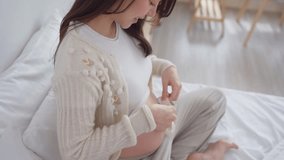 Pregnant woman gently places baby shoes on belly while sitting on bed. Attractive expectant mother feel happy and relax, preparing for maternity and embracing beauty of pregnancy in bedroom at home. - Powered by Shutterstock - Get 15% off with code: PIKWIZARD15