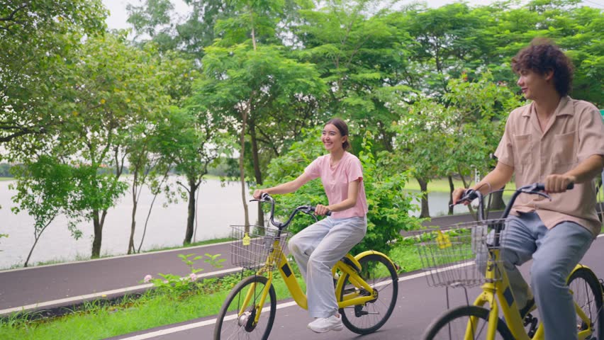 Asian young couple dating and biking together on a road in the park. Attractive man and woman feel happy and enjoy riding bicycles side by side, spend free leisure time and outdoors activity in garden