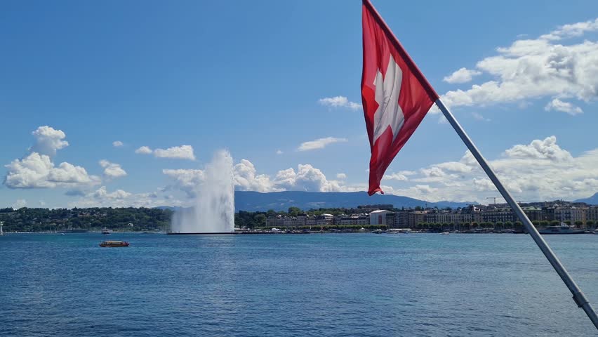 Swiss flag waving in the wind with Lake Geneva in the background. The famous Jet d’Eau fountain is visible in the distance under a bright blue sky with clouds. A sunny summer day in Geneva,Switzerland