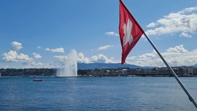 Swiss flag waving in the wind with Lake Geneva in the background. The famous Jet d’Eau fountain is visible in the distance under a bright blue sky with clouds. A sunny summer day in Geneva,Switzerland - Powered by Shutterstock - Get 15% off with code: PIKWIZARD15