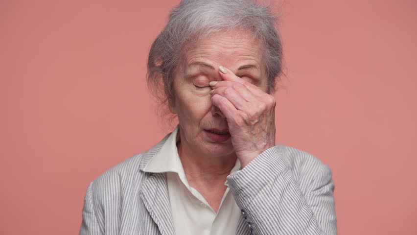 Senior woman in striped jacket holds her forehead and closes her eyes, showing pain and discomfort from a headache, isolated on pink background.