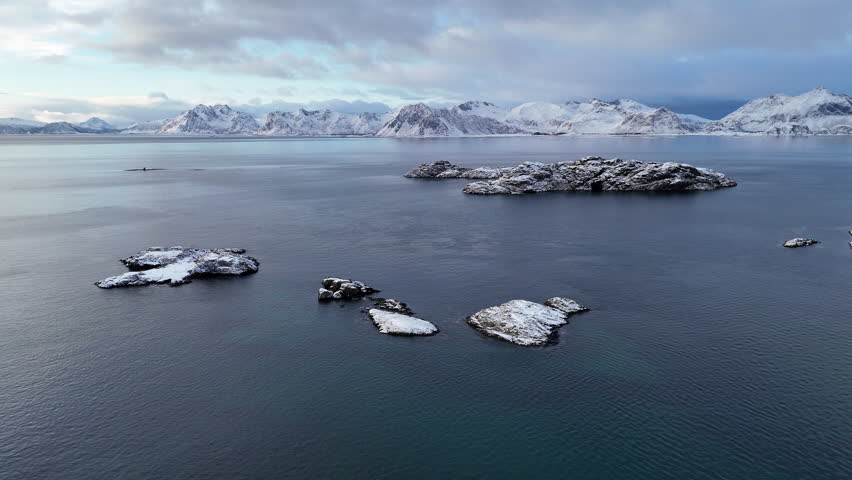 Aerial view of snow-covered islets in the Arctic Ocean near Lofoten, Norway, with dramatic mountain backdrop, serene winter seascape, and breathtaking Nordic coastal scenery