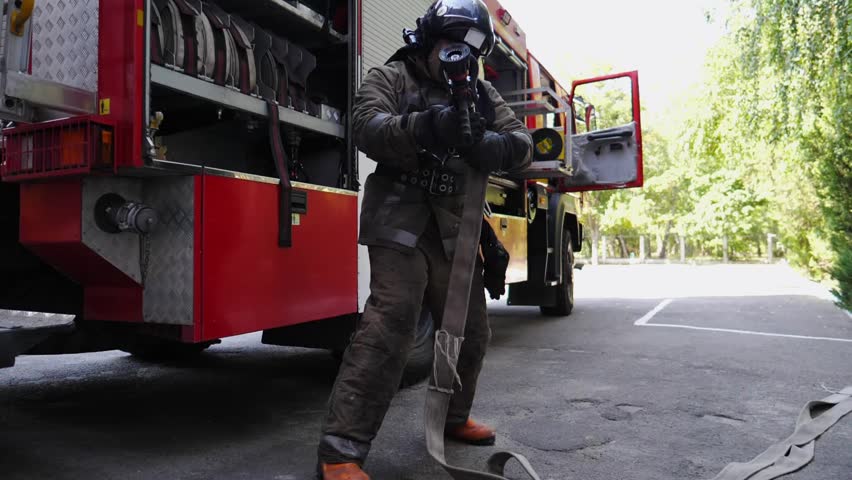 Professional fireman in full equipment connecting fire hydrant to hose near big red truck. Young male fireguard getting ready to extinguish fire. Saving lives and heroic profession concept. Slow mo