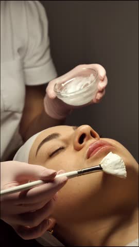 A beautician applies a white cleansing mask to a female face close-up, vertically. A woman at a relaxing spa treatment enjoys