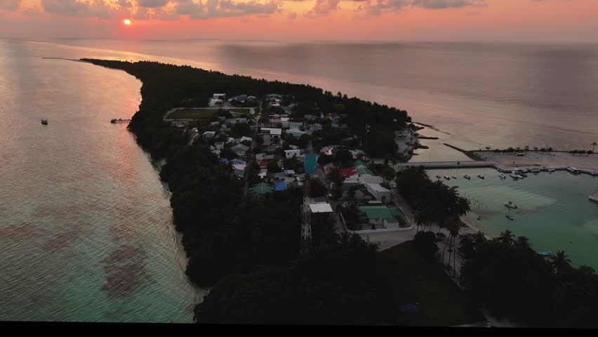Aerial view of Fulhadhoo Island showcasing the tranquil village, lush green trees, and turquoise waters under the soft glow of the setting sun, Fulhadhoo, Baa Atoll, Maldives.