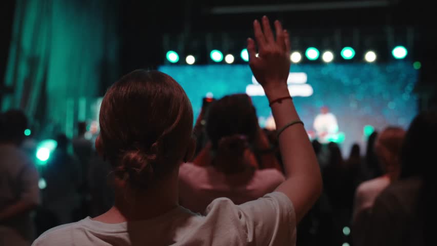 Female worshipper lifting hand during live music event, surrounded by crowd, large screen projecting colorful background visuals