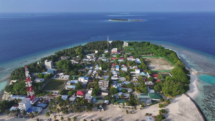 Aerial view of turquoise waters meeting the sandy shores of Kamadhoo, with buildings nestled amongst lush greenery, Kamadhoo, Baa Atoll, Maldives.
