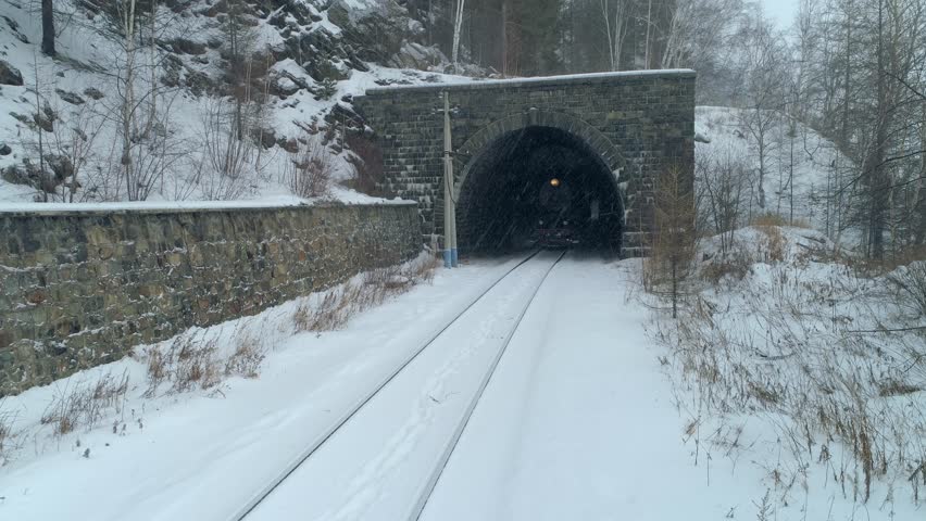 Flight upwards old steam locomotive emerging from tunnel with steam plume from chimney. Unique winter footage of historical northern railway in snowstorm blizzard conditions, atmospheric scene 4k