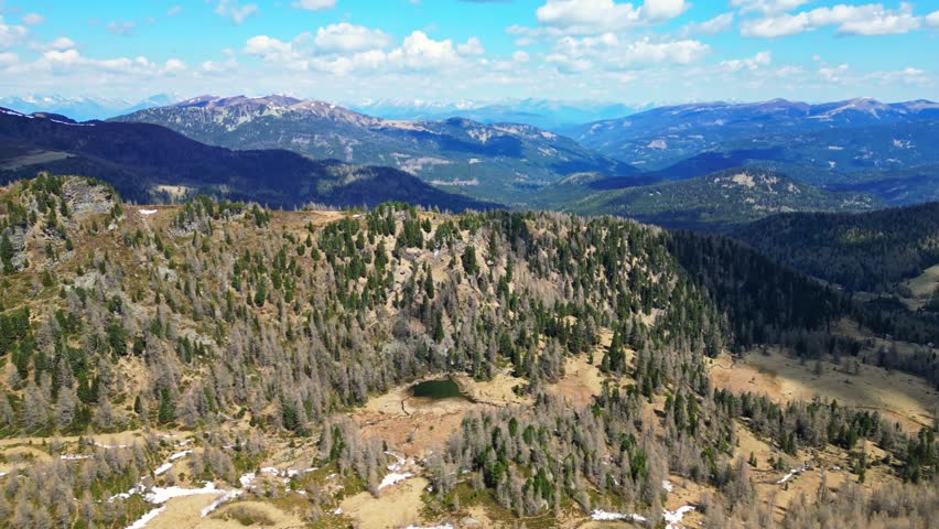 Aerial view of hills displaying a dense forest with a small lake, under a blue sky dotted with fluffy clouds, Hochrindl, Kärnten, Austria.