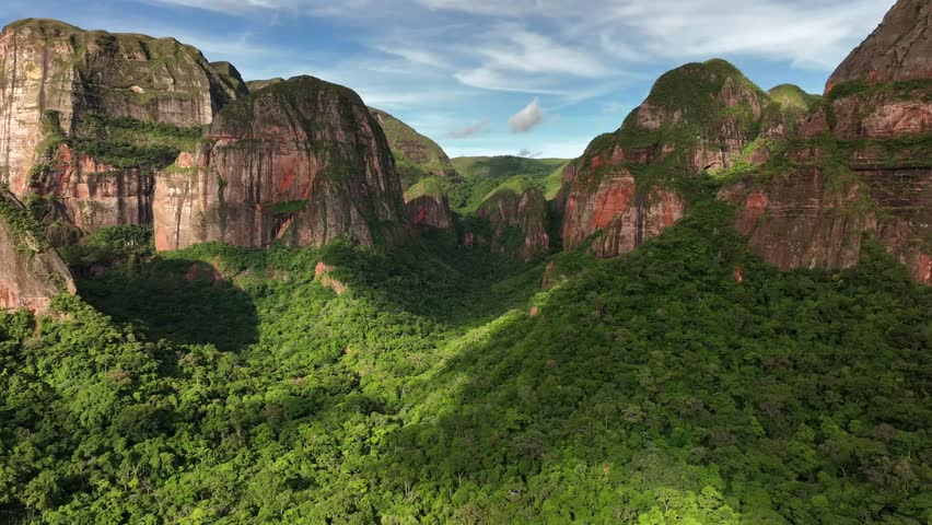 Aerial view of lush green forests meeting the red rock mountains under a blue sky with scattered clouds, Amboro national park, Samaipata, Bolivia.