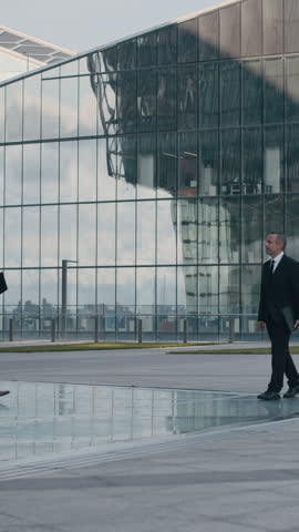 Vertical shot of male middle-aged entrepreneurs sharing document when meeting on city square with glass buildings