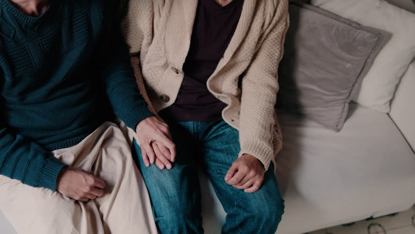 A gay couple sits together on a sofa, holding hands and sharing an intimate moment of affection. This clip portrays a sense of love, comfort, and togetherness.