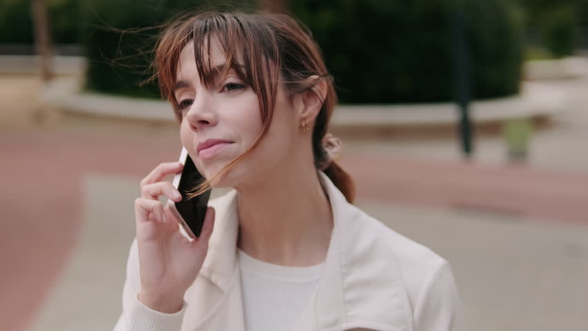 A young woman smiles while talking on her phone outdoors. She walks along a brick pathway past some trees and shrubbery.