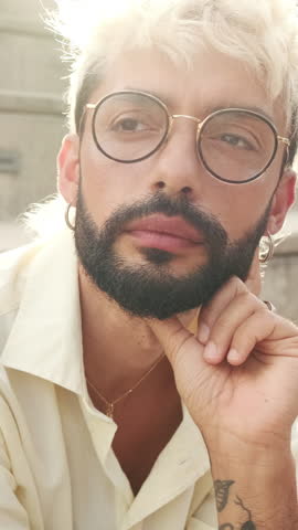 Vertical video, Portrait of a thoughtful young man with bleached blond hair and a beard, wearing glasses and a cream shirt, sitting on steps, looking away.