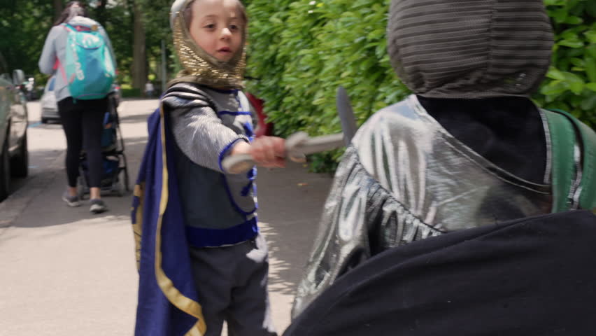 Child dressed as a knight swings a toy sword in a lively street battle with a friend. The playful moment captures the thrill and imagination of children engaging in outdoor role-play adventures
